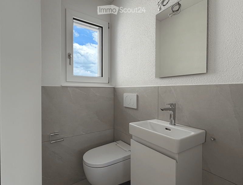 Bathroom. Modern bathroom with a white sink, chrome faucet, toilet, and large mirror above the sink. Grey tiled walls and a window with a view of the sky.