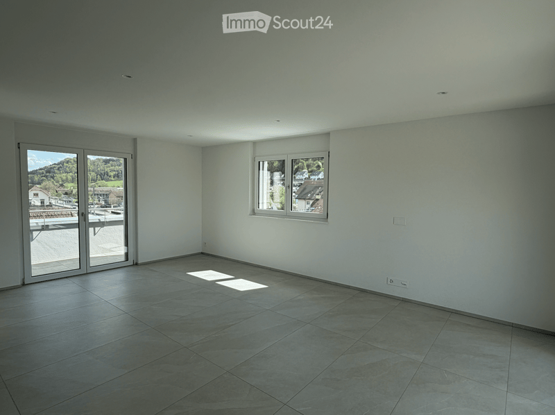 Unfurnished Room. Empty room with light grey walls, tiled floor, large sliding glass door leading to a balcony, and a window with a view of the town and surrounding landscape.