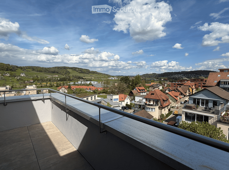 Balcony / Terrace. Balcony with metal railing overlooking a town with houses, green fields, and hills under a partly cloudy sky.