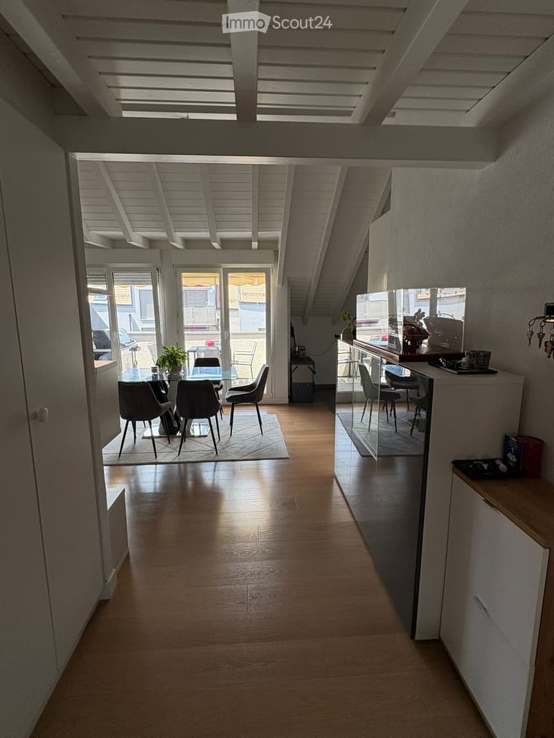 Bright, open corridor with wooden flooring leading to a dining area and kitchen. The corridor has white walls and a slanted ceiling with exposed beams. The dining area features a glass table, four chairs, and a small plant. The kitchen area includes a cou