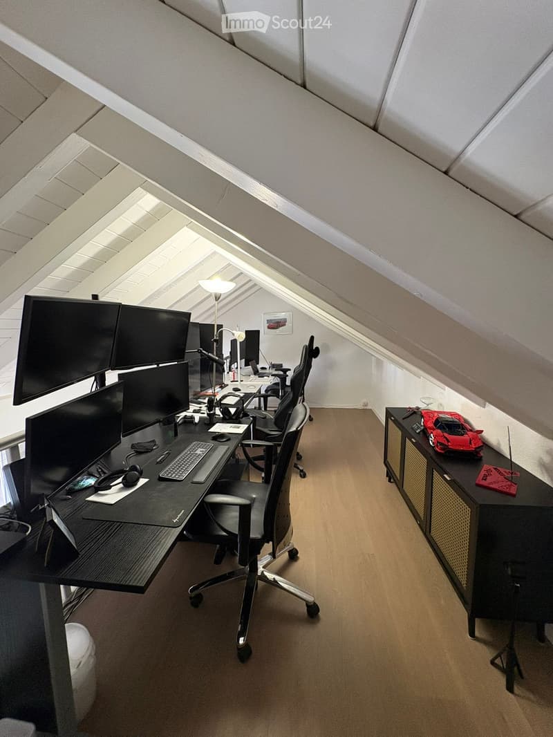 Modern attic office with slanted ceilings, multiple computer screens, ergonomic chair, desk, and a small cabinet with a toy car on top.