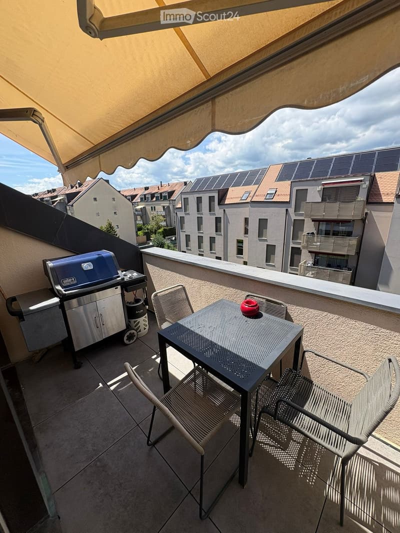 Balcony with a table, chairs, and a grill, featuring a retractable awning and a view of neighboring buildings.