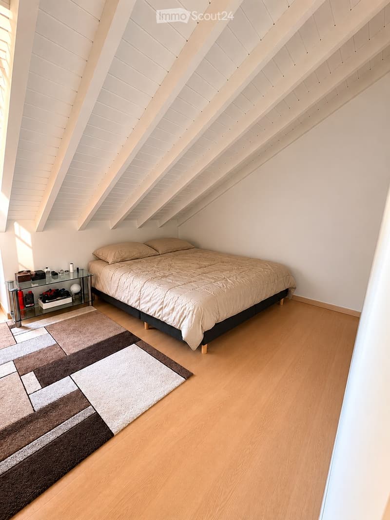 Bright attic bedroom with wooden slanted ceilings, light wooden flooring, a double bed with beige bedding, a nightstand with a mirror, and a patterned rug.