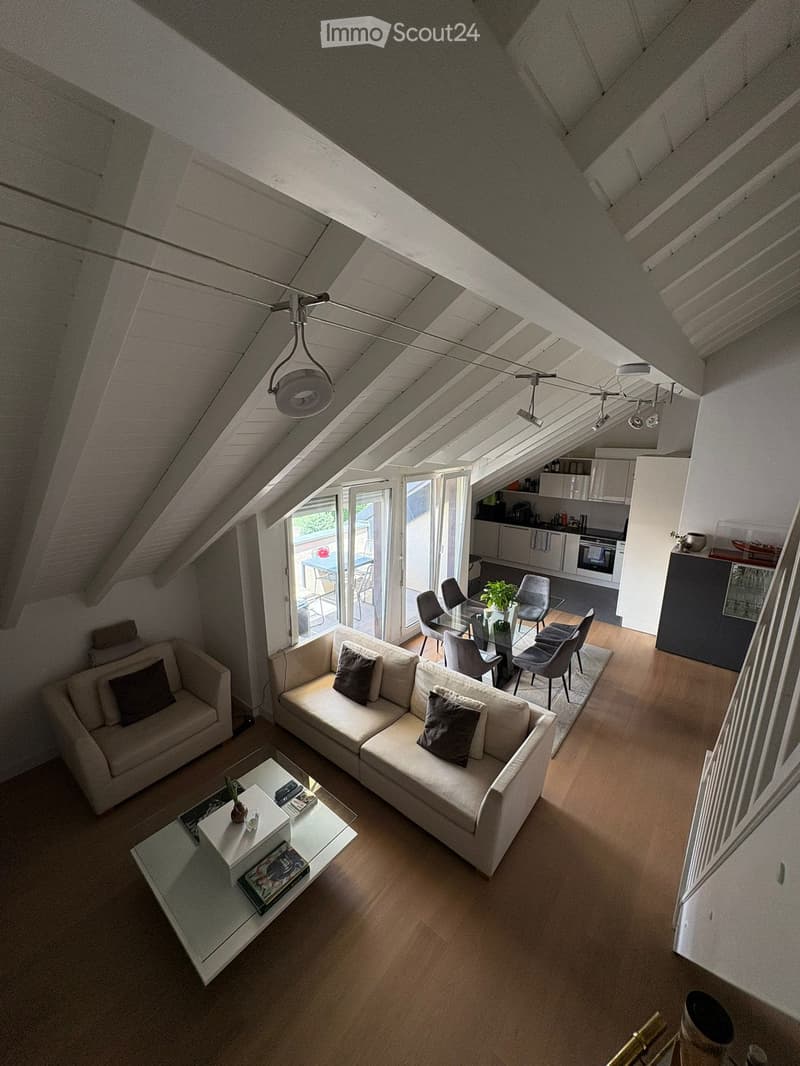 Bright, open living space with wooden flooring, white walls, and exposed wooden beams. Features a large sectional sofa, coffee table, dining area with chairs and table, and a modern kitchen with cabinets and appliances.