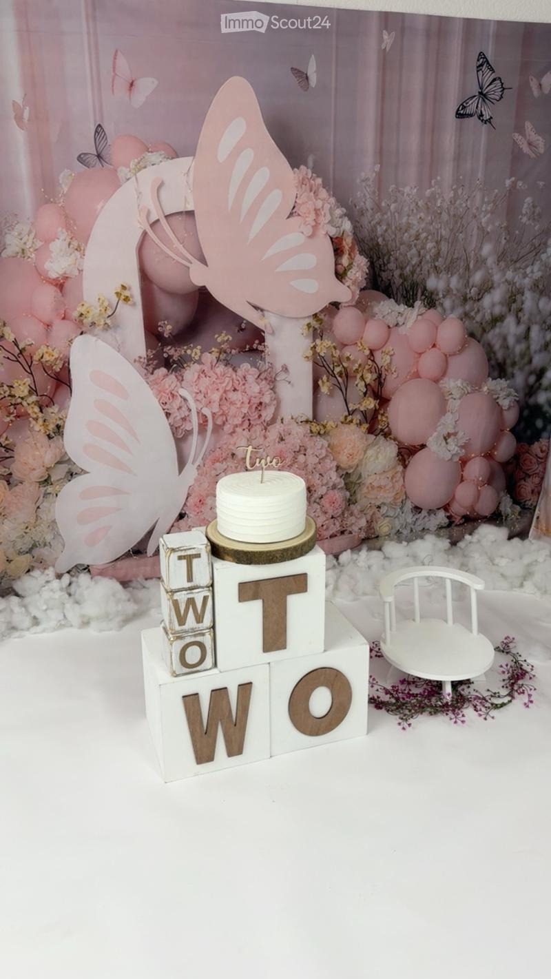 A decorative setup featuring a white cake with a candle, pink balloons, floral arrangements, butterfly decorations, and a small white chair.