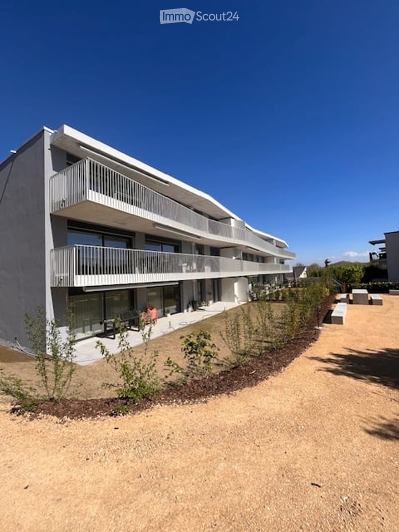 Modern multi-story building with white balconies and large windows, surrounded by small plants and a sandy pathway.
