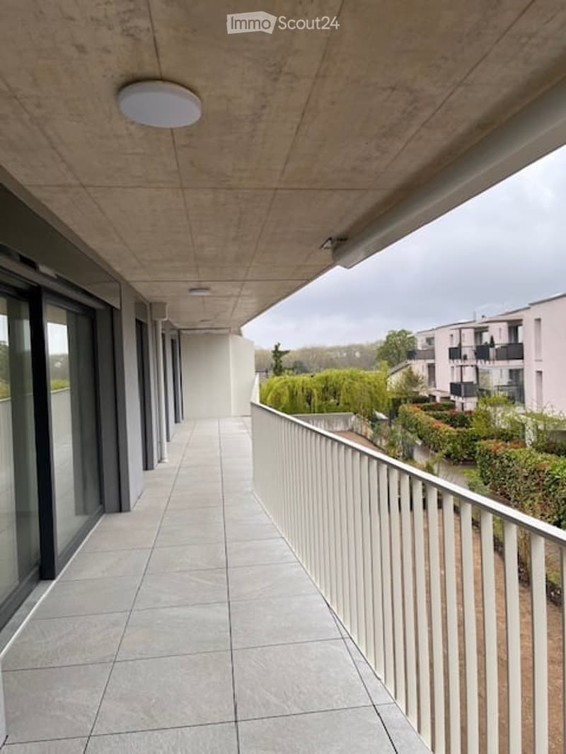 Modern balcony with concrete floor and ceiling, white railing, large glass windows, and a view of greenery and neighboring buildings.