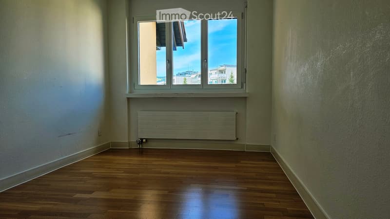 Empty room with wooden flooring, white walls, and a large window offering a view of the outside. A radiator is mounted below the window.