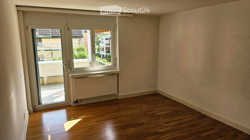 Empty room with wooden flooring, large window with blinds, radiator, and a view of the balcony and surrounding buildings.