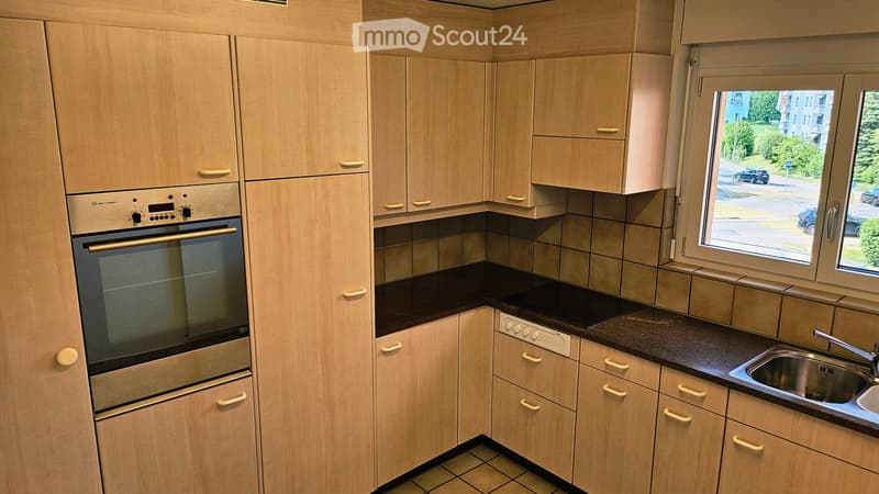 Modern kitchen with light wood cabinets, stainless steel oven, and a window overlooking the street.