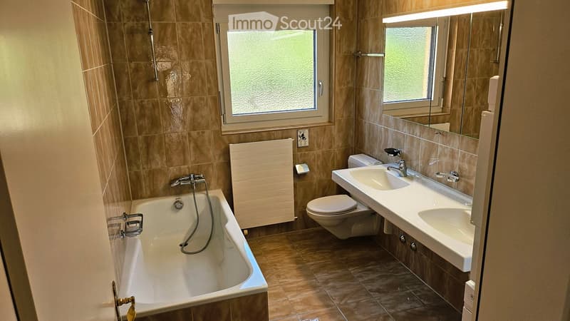 Modern bathroom with beige tiles, a bathtub, a toilet, a double sink, a large mirror, and a radiator.