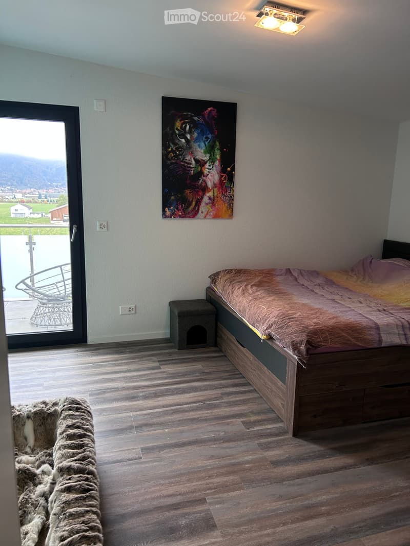 Modern bedroom with wooden flooring, a large bed with a fur throw, a small stool, a chair, and a colorful painting on the wall. A sliding glass door leads to a balcony with a scenic view.
