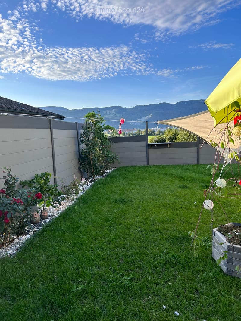 Well-maintained garden with green grass, small plants, and a decorative fence, featuring a view of distant mountains.