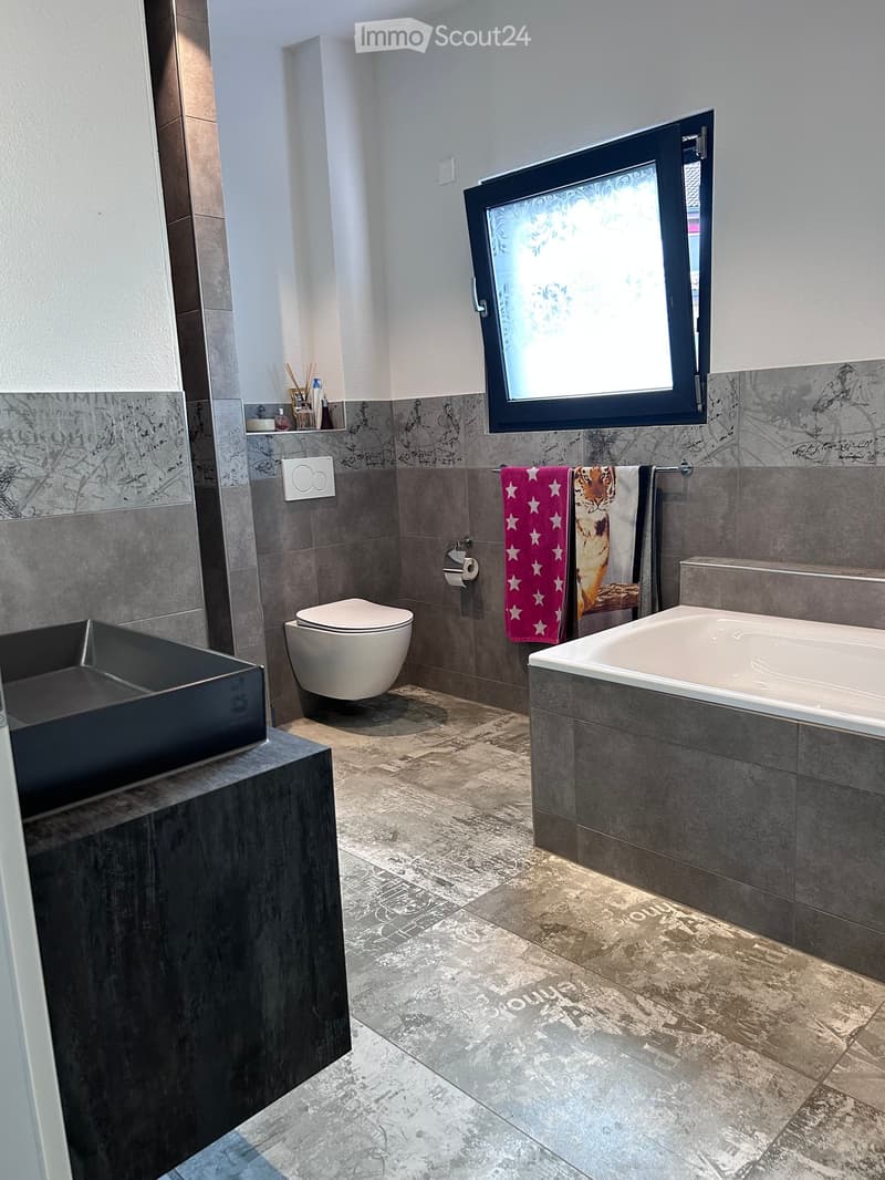Modern bathroom with a bathtub, toilet, sink, and towels. Features grey tiles, a window with frosted glass, and a dark wooden vanity.