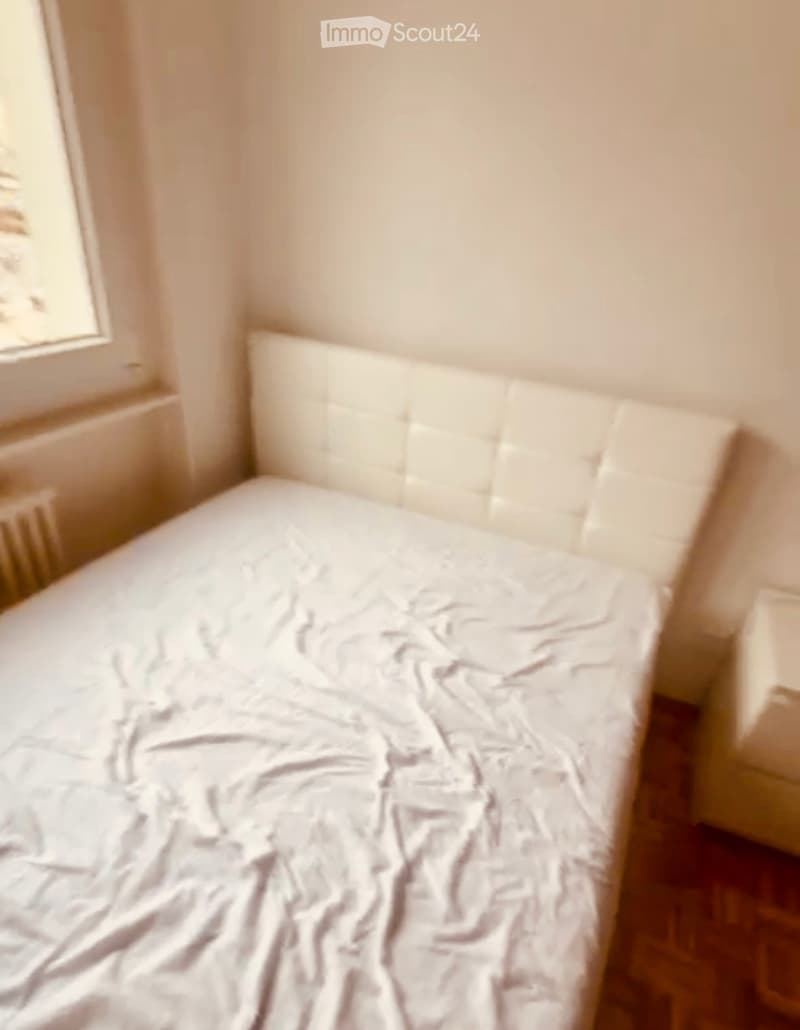 A small bedroom with a single bed, white headboard, and a window on the left side.