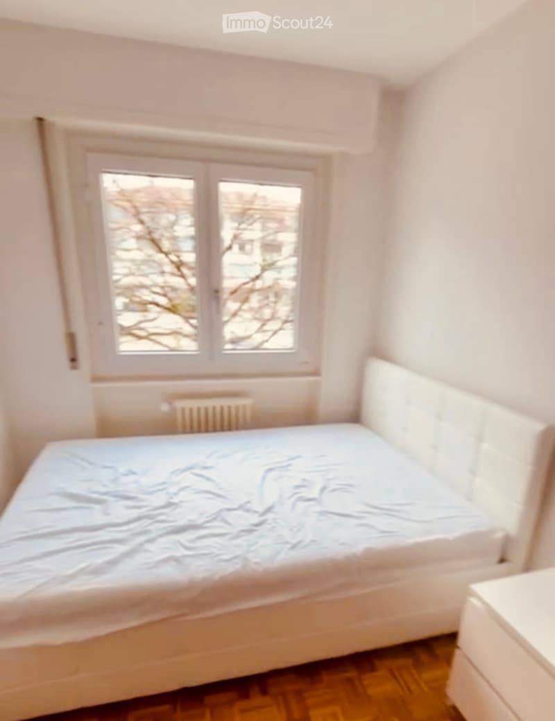 A small bedroom with a single bed, wooden flooring, and a window with a view of trees. A radiator is placed below the window.