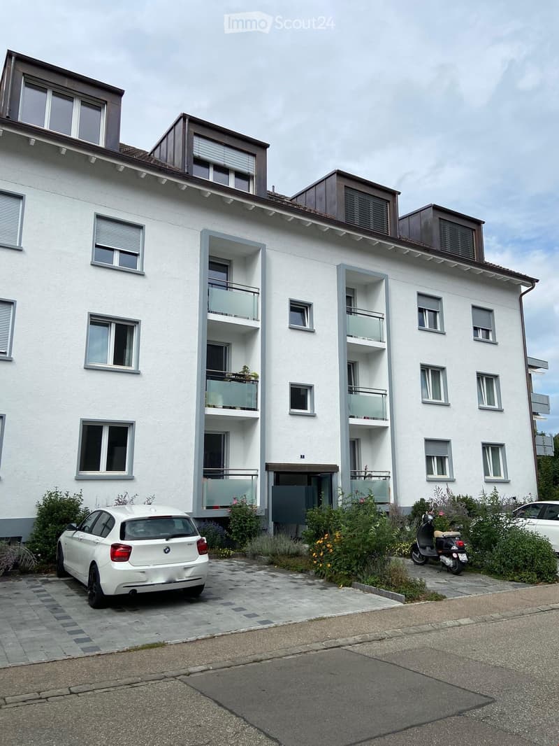 Modern three-story apartment building with white facade, multiple windows, and balconies. A white BMW car and a scooter are parked in the driveway.