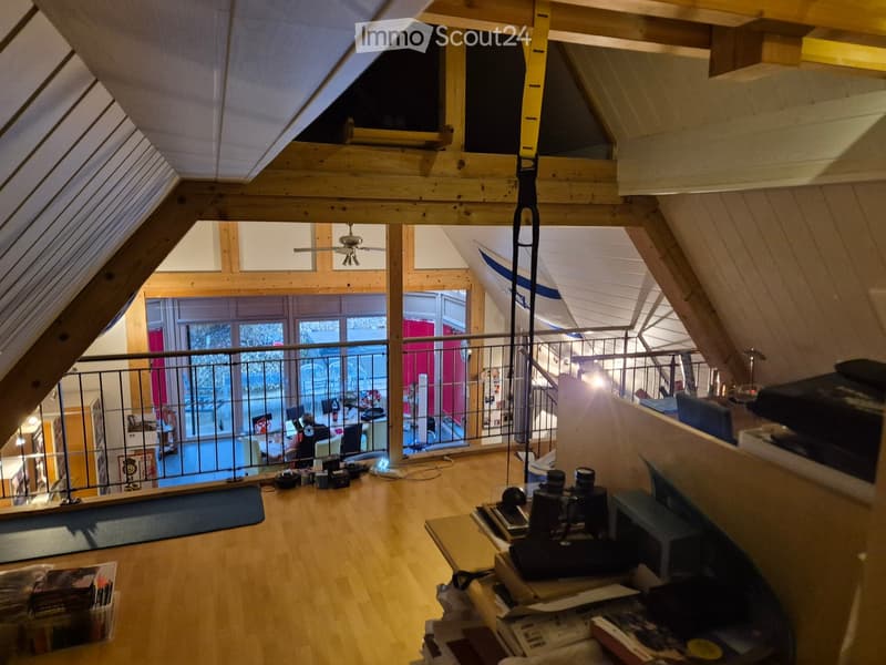 Loft-style room with wooden floors, slanted ceilings, and large windows overlooking a lower level area. Visible items include a table with a laptop, chairs, books, and binoculars.