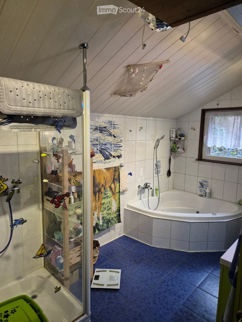 Cozy attic bathroom with a bathtub, shower, towel, shelf, and decorative elements, including a fish-themed design on the tiles and a small window with a view.