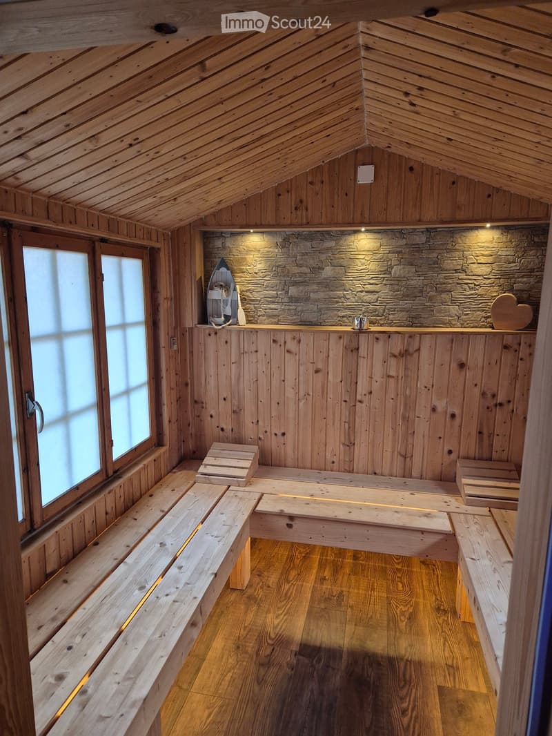 Cozy wooden sauna room with slanted wooden ceiling, wooden benches, small stools, and a stone accent wall with a boat model and heart-shaped decoration.