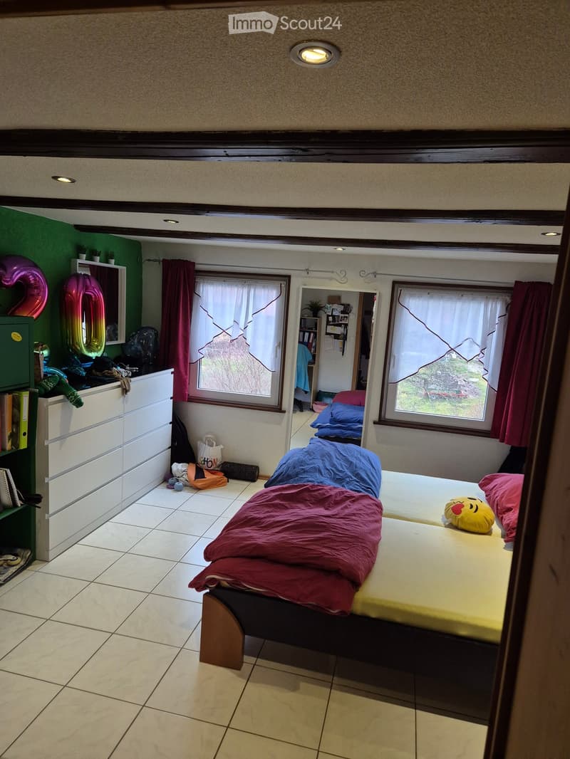 Cozy bedroom with a bed covered in blankets and pillows, a bookshelf, a mirror, and balloons. The room features tiled flooring, large windows with curtains, and a small closet area visible in the background.