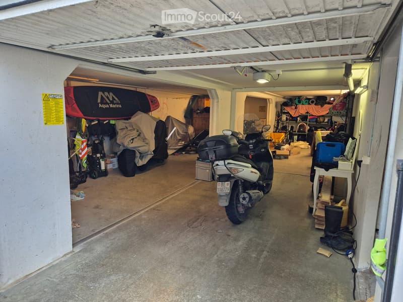 Underground garage with concrete floor, white ceiling beams, and storage items including a scooter, covered furniture, and various equipment.