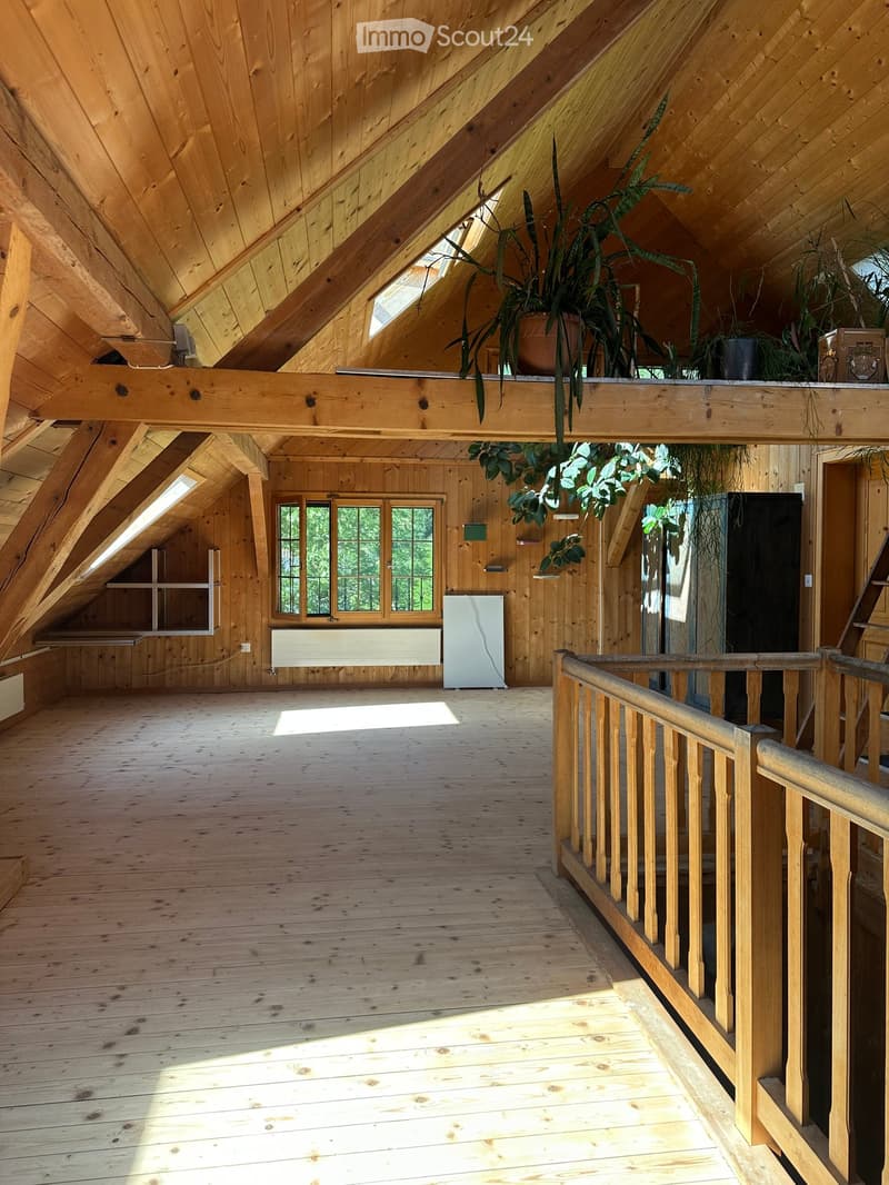 Spacious attic room with wooden beams, wooden floor, large windows, and a balcony area with wooden railing and potted plants.