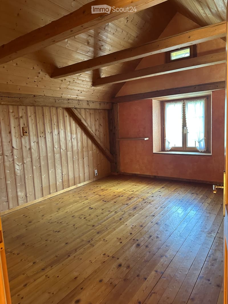 Empty attic space with wooden walls, wooden floor, exposed wooden beams, and a small window with curtains.