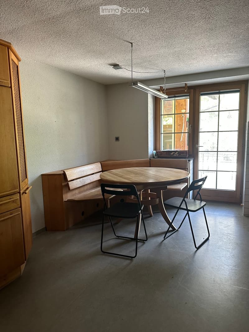 A small, modern dining area with a round wooden table, two folding chairs, and a built-in wooden bench seating along one wall. The room has large windows and a sliding glass door leading outside.