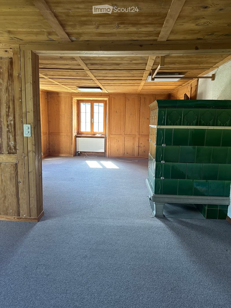Empty room with wooden walls, exposed wooden beams on the ceiling, carpeted floor, and a radiator near a window. A green fireplace is visible on the right side.