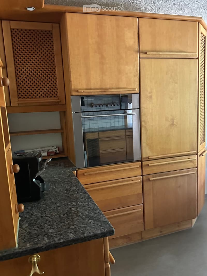 A modern kitchen with light wooden cabinets, a granite countertop, and a built-in oven.