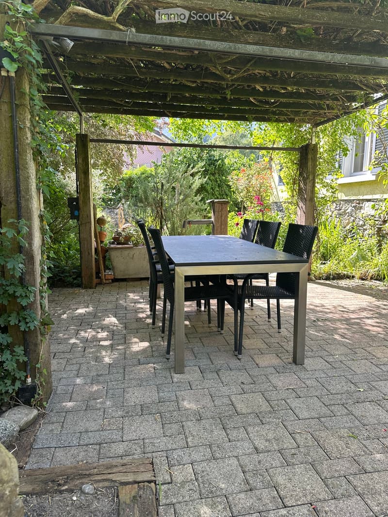 A covered outdoor patio with a stone-tiled floor, a black metal table surrounded by black wicker chairs, and lush greenery in the background.