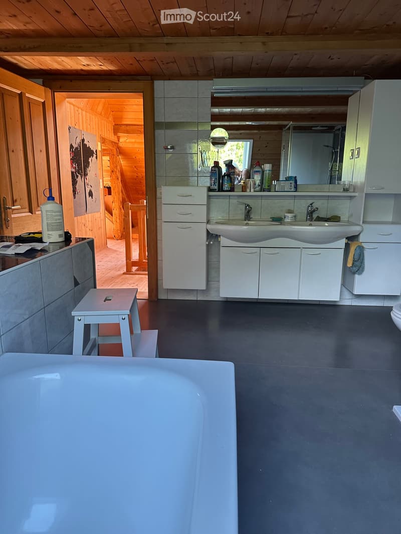 Modern bathroom with a double sink, white cabinets, a large mirror, a bathtub, and a small white stool. The room features grey tiles and a wooden ceiling.
