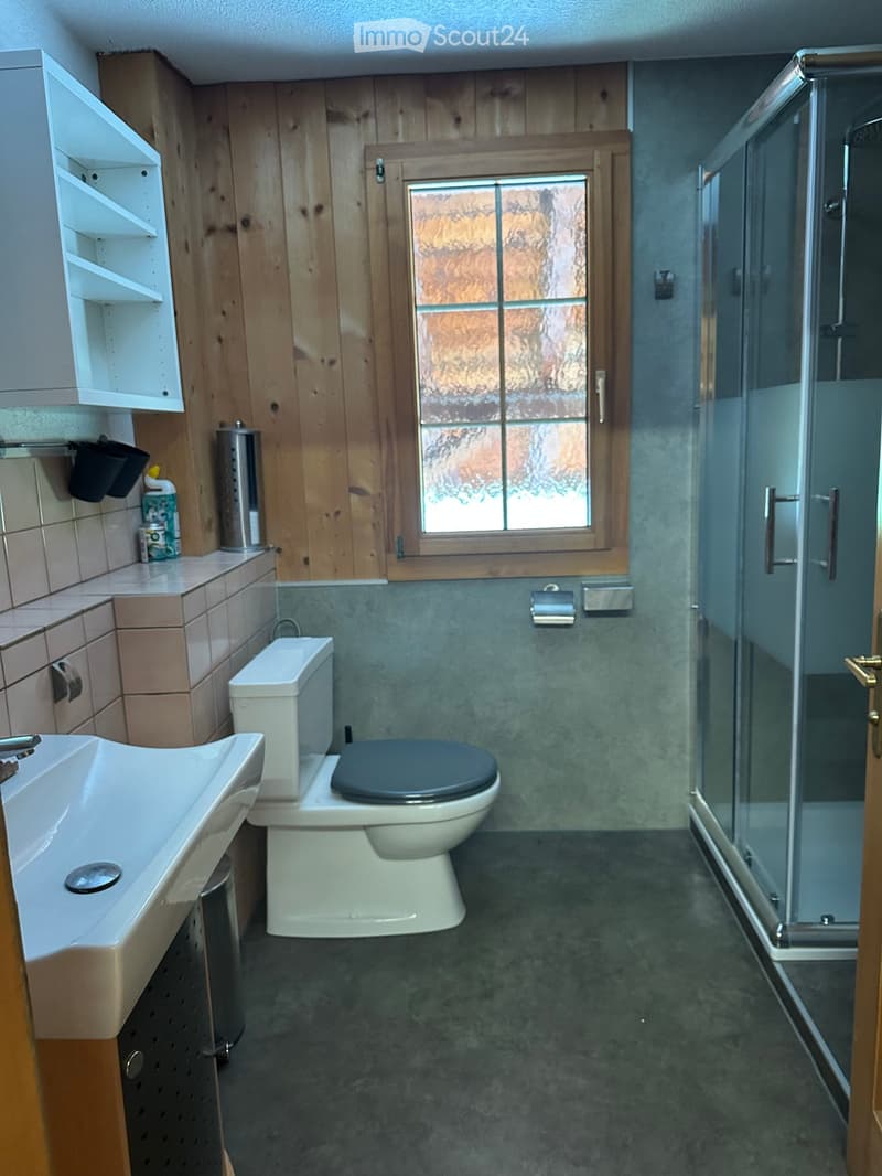 Bathroom with a sink, toilet, shower, and wooden cabinetry. Features a window with frosted glass and a tiled floor.