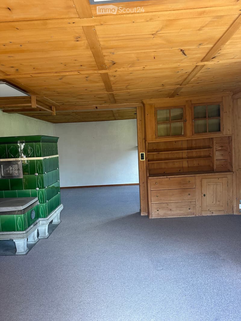 Empty room with wooden ceiling beams, blue carpet, green tiled stove, wooden cabinet with glass doors, and a small window.