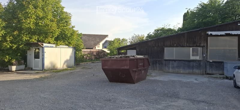 Outdoor area with a large wooden shed, a red dumpster, a small white trailer, and a parked car. Trees and a neighboring house are visible in the background.