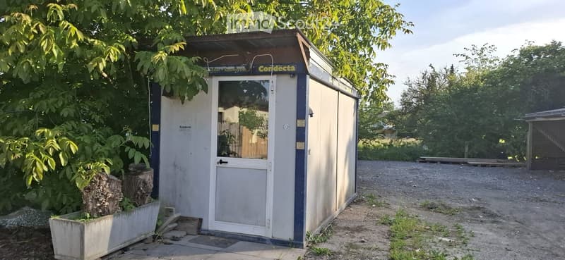 A small, portable shed with a white and blue exterior, situated in a garden area with trees and a gravel path.