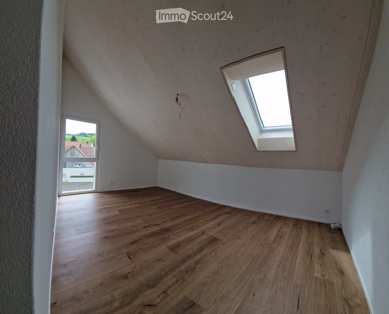 Empty room with wooden floors, white walls, and two windows. One window has a view of a house and trees outside.