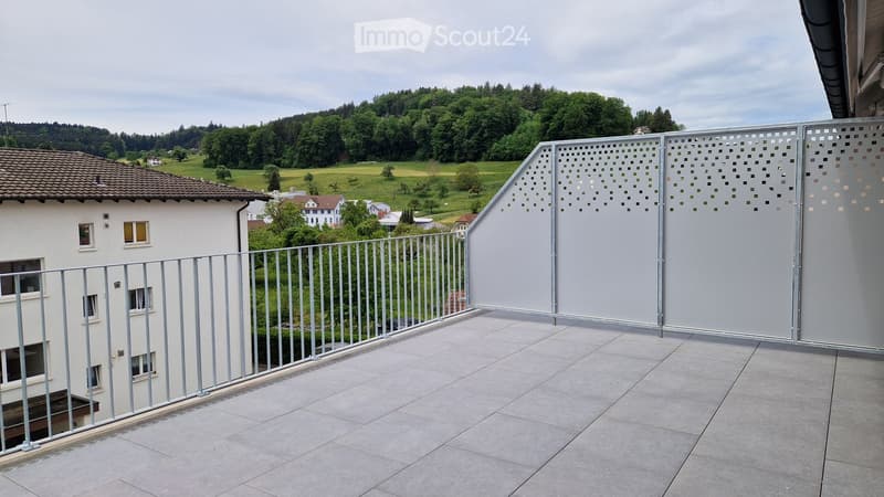 A balcony with a metal railing and a decorative metal fence. The floor is tiled. The view shows a green landscape with houses and trees.