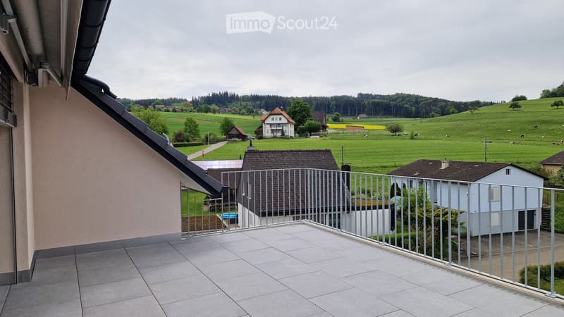 A balcony with a metal railing and tiled floor, overlooking a rural landscape with houses and fields.