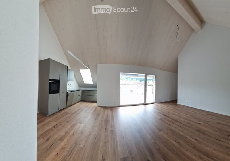 An empty room with wooden floors, white walls, and a ceiling with wooden beams. There is a kitchen area with cabinets, a sink, and a refrigerator. There is a glass door leading to a balcony with a view of the outside.
