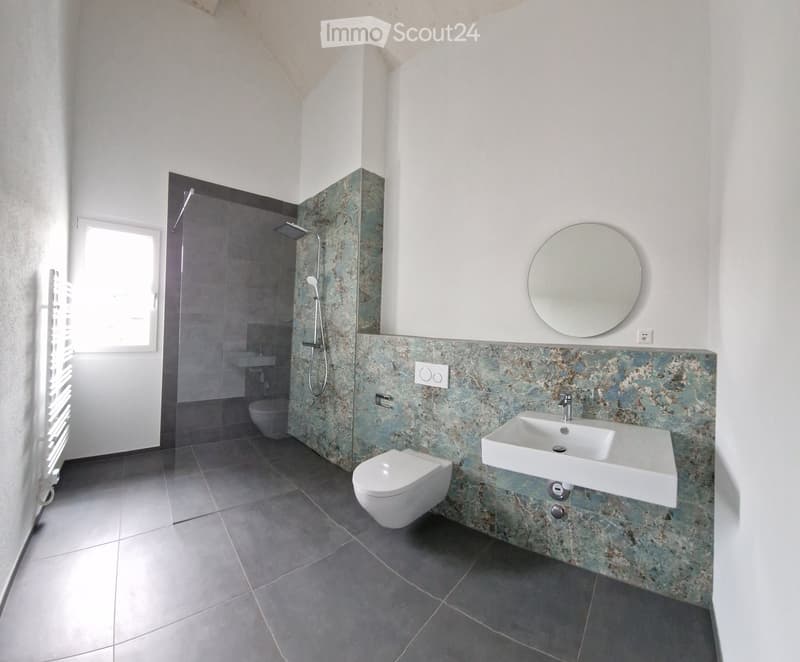 Modern bathroom with gray tiles, white toilet, shower, sink, and round mirror.