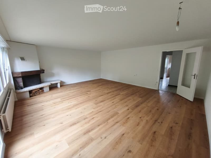 Empty room with wooden flooring, white walls, and a fireplace with a bench and wood stack. A doorway leads to another room.