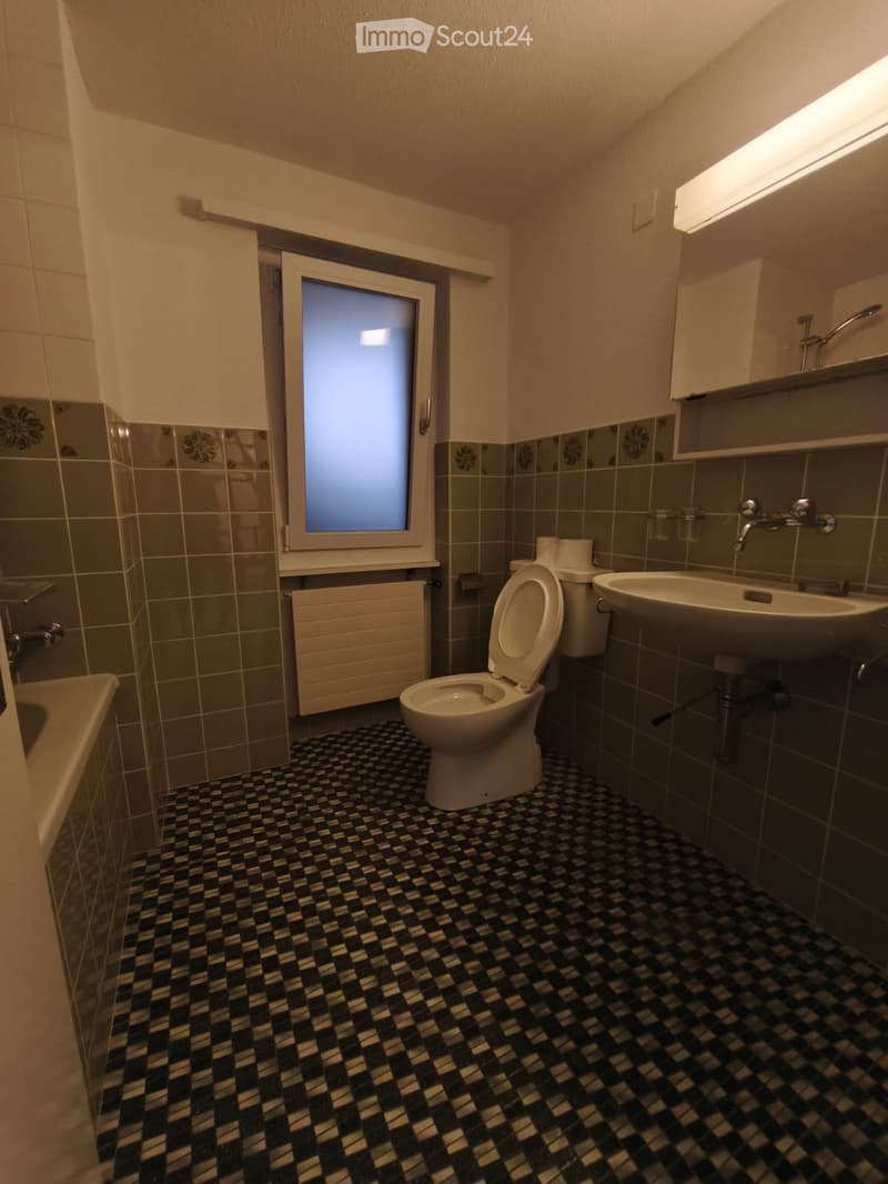 Bathroom with checkered black and white floor tiles, grey tiled walls, a toilet, a sink, a bathtub, and a window with frosted glass.