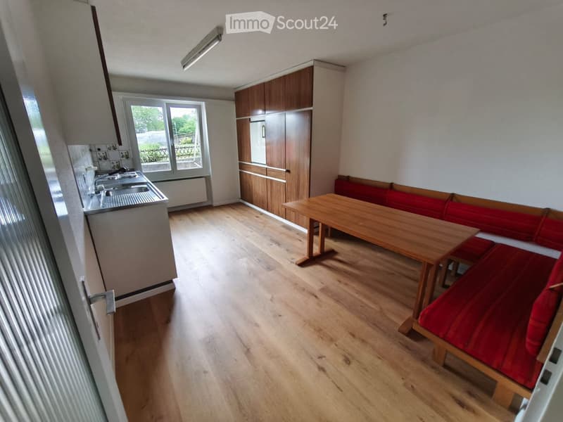 A compact, well-lit room with wooden flooring, a wooden table, red bench seating, and a small kitchen area with cabinets and a sink.
