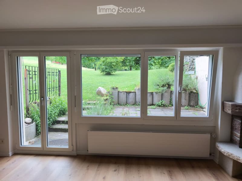 Empty room with wooden flooring, large windows, radiator, and sliding glass doors opening to a garden.