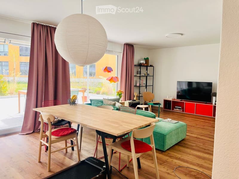 Spacious room with wooden floor, dining table, and chairs. Green couch with pillows, TV on a shelf, and a treadmill. A lamp and a plant add to the decor.