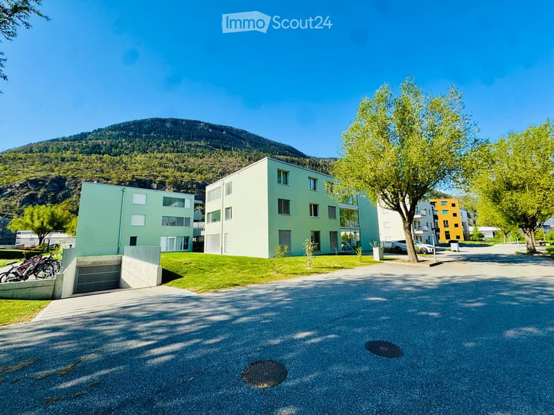 Modern apartment buildings with green facades, surrounded by greenery and a mountain in the background.