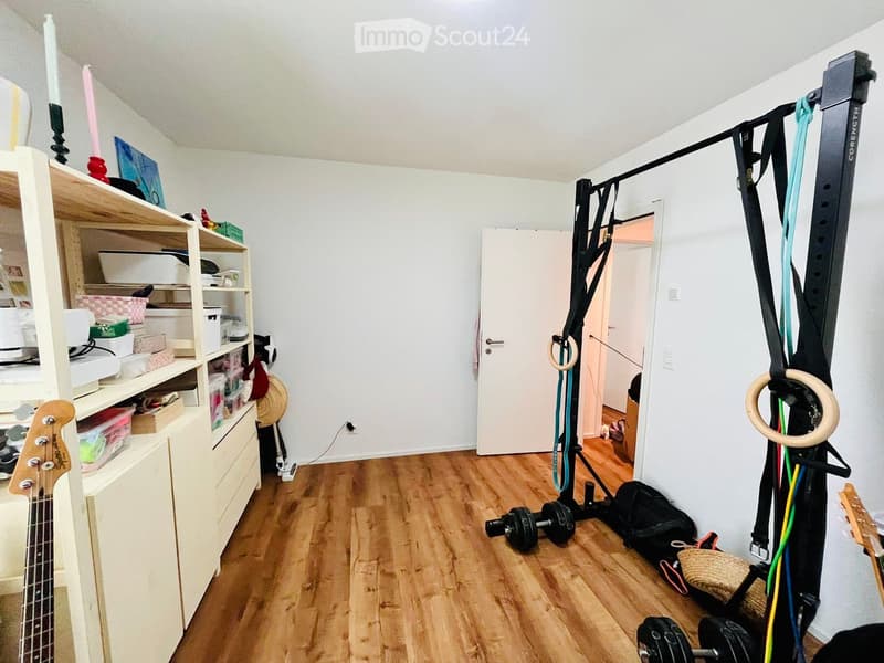 A room with wooden floor, white walls, and a white door. There is a shelf with various items, a guitar, dumbbells, a dumbbell rack, and a basket on the floor.
