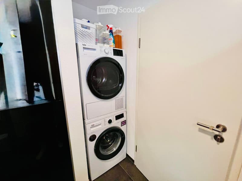A small room with a white door, a white washing machine, and a dryer stacked on top of each other. There is a basket on top of the dryer and cleaning products on the shelf above the machines.
